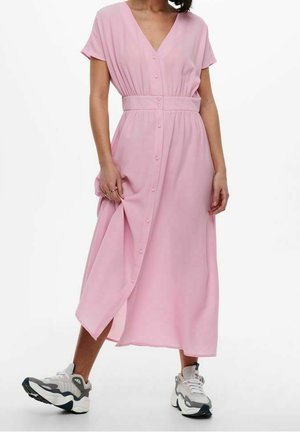 Kobieta ubrana w jasnoróżową sukienkę na guziki z paskiem, w połączeniu z masywnymi białymi i szarymi trampkami, stojąca na białym tle.