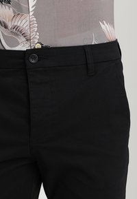 Černé kalhoty vyrobené z hladké tkaniny, s jednořadovým zapínáním na knoflík, poutky na opasek a šikmými bočními kapsami.