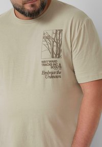 Behaarde man die een beige T-shirt draagt met een bruine boomafbeelding en de tekst "Wayward Tracks Inc. & Scouts" en "Omarm het Onbekende" op de borst.