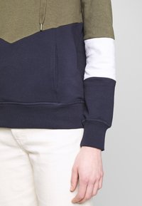 Sudadera con bloques de color que presenta paneles en verde olivo, azul marino y blanco, hecha de una tela suave con capucha ajustable y puños de canalé.
