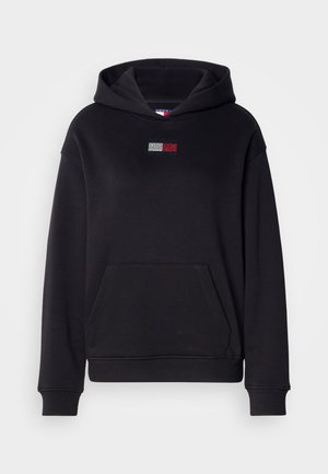 Čierna mikina s kapucňou a predným vreckom, ktorá má na hrudi malé, farebné logo vyrobené z kamienkov. Vyrobená z mäkkej, texturovanej látky.