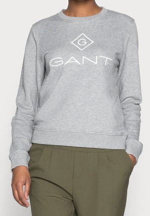 Personne portant un sweat-shirt GANT gris clair à manches longues et un pantalon vert olive, debout avec une main dans la poche.