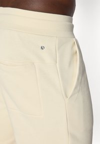 Cremfarbene Jogginghosen mit weicher Textur, ausgestattet mit einem gerippten Taillenbund, Seitentaschen und einem kleinen gestickten Logo nahe der Tasche.
