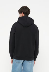 Hoodie preto com um bolso canguru e capuz ajustável com cordão. Tecido suave, punhos e barra canelados, usado com jeans baggy azul claro.