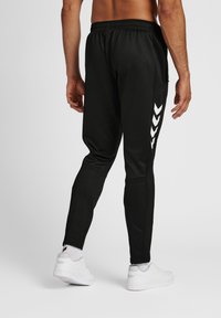 Svarta träningsbyxor med slim fit, med ett vitt chevronmönster på vänster sidan och en elastisk midja. Vita sneakers bärs.
