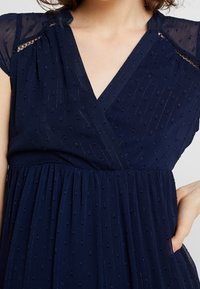 Detailní záběr na námořnické šaty s texturovanými tečkami, průsvitnými krátkými rukávy, výstřihem do V a rukou odpočívající na postranní kapse.