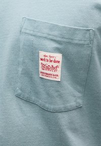 Φωτεινό γαλάζιο ύφασμα τσέπης πουκαμίσου με λευκή ετικέτα στην οποία αναγράφεται με κόκκινα γράμματα «όταν υπάρχει δουλειά να γίνει» και κόκκινο λογότυπο με άμαξα που την τραβά άλογο.