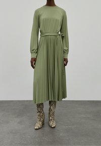 Grön veckad klänning med långa ärmar och knutet midjeband, kombinerad med knähöga stövlar med ormskinnsmönster. Slät, lätt tyg.