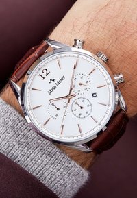 White chronograph watch with silver accents, brown leather strap, and three sub-dials. Hour markers in rose gold. Stainless steel case.