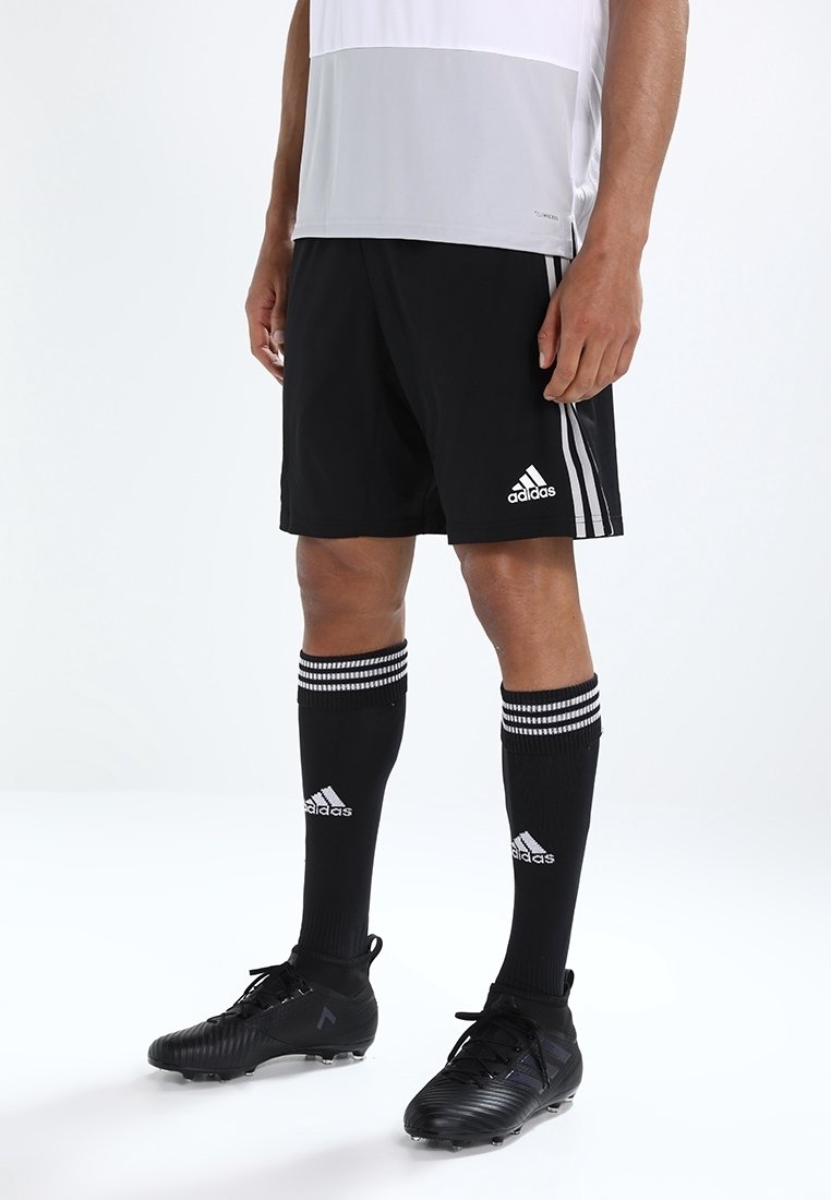 Muž v černých fotbalových šortkách Adidas, černých podkolenkách Adidas a černých kopačkách, stojící na bílém pozadí.