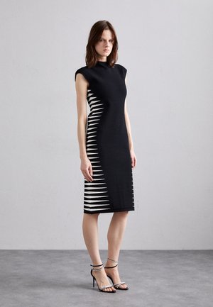 Abito nero al ginocchio con maniche a sbuffo, caratterizzato da un motivo a zigzag bianco sul lato. Tessuto morbido con una silhouette aderente. Tacchi neri con cinturini.