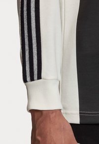 Hvid sportslig sweaterærme med sort og grå tre-stribe design. Ribbet manchetter og glat tekstur. Nærbillede af hånd, der hviler i nærheden.