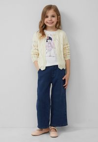 T-shirt blanche avec un personnage imprimé, surmontée d'un cardigan en tricot crème. Jean large en denim et chaussures Mary Jane rose pâle.