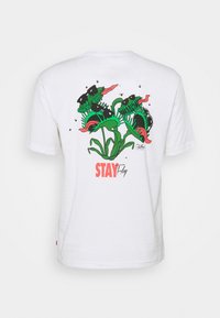 Valkoinen puuvillapaita, jossa on värikäs grafiikka kahdesta lihansyöjäkasvista ja teksti "STAY Fly" kirkkaan punaisena. Rentoa istuvuutta ja lyhyet hihat.