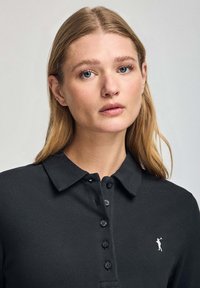 Polo negro con tejido texturizado, cuello corto y tapeta de tres botones. Presenta un pequeño logo blanco en el lado izquierdo.