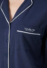 Camisa de pijama azul marinho feita de tecido macio, com acabamento em branco, fechamento por botão e um bolso à esquerda no peito com bordado do logotipo.