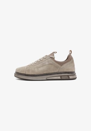 Zapatilla de ante beige con punta redondeada, cordones planos y suela acolchada. Presenta un talón en contraste marrón y acentos de costura blanca.