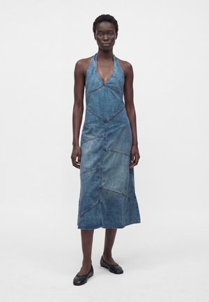 Femeie purtând o rochie halter din denim albastru fără mâneci cu cusături patchwork și pantofi negri comozi, stând pe un fundal alb simplu.