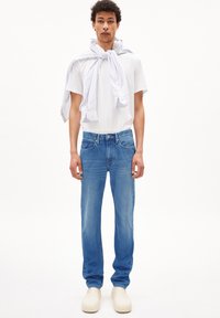 T-shirt blanc léger, jean droit bleu moyen et sabots beiges. Chemise rayée bleue et blanche nouée autour du cou.