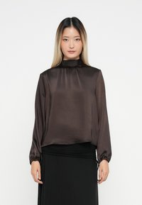 Woman with long ombre hair wearing a dark brown satin high-neck blouse and a black skirt, standing against a white background.