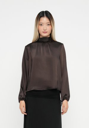 Woman with long ombre hair wearing a dark brown satin high-neck blouse and a black skirt, standing against a white background.