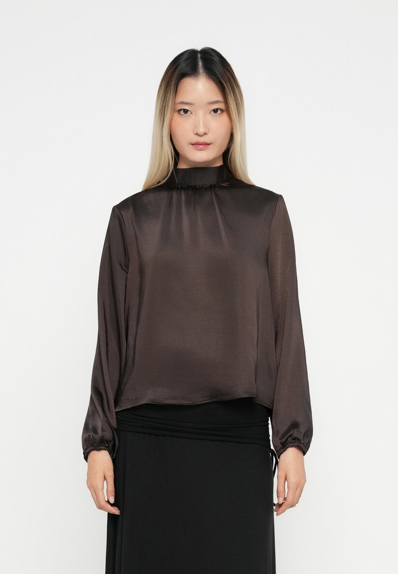 Woman with long ombre hair wearing a dark brown satin high-neck blouse and a black skirt, standing against a white background.