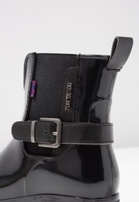 Botín negro con acabado brillante de charol, paneles laterales de tela, detalle de hebilla de cinturón y una pequeña etiqueta de logo en colores contrastantes.