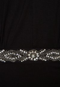 Vêtement en tissu noir présentant un ornement en perles avec des strass clairs et argentés disposés selon un motif décoratif à la taille.