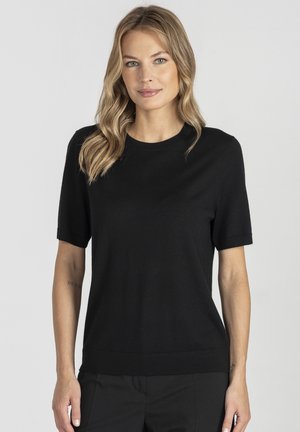 Blondynka z falującymi włosami w czarnej bluzce z krótkim rękawem i czarnych spodniach, stojąca na tle jasnego, jednolitego tła.