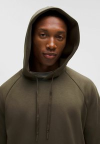 Oliwkowa, kapturowa bluza o gładkiej teksturze, z akcentami w postaci sznurka i rękawami typu raglan. Minimalistyczny design, bez widocznych logotypów czy wzorów.