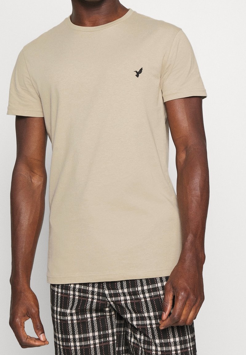 Homme portant un t-shirt beige à manches courtes avec un petit logo d'oiseau noir sur la poitrine et un pantalon à carreaux noir, blanc et marron, sur fond blanc.