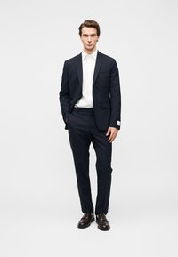 Σκούρο μπλε κοστούμι, εφαρμοστή γραμμή με δύο κουμπιά, μία τσέπη στο στήθος. Λευκό πουκάμισο με κουμπιά από κάτω, σε συνδυασμό με καφέ παπούτσια.