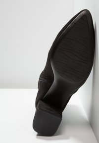 Botte noire en daim à bout pointu avec semelle en caoutchouc et petit talon bloc, vue de dessous sur fond blanc.