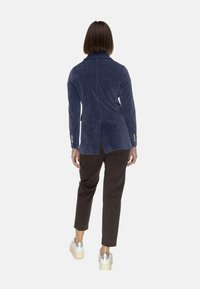 Blazer in velluto blu navy con spacco posteriore e dettagli in bottone dorato, abbinato a pantaloni marroni e scarpe con plateau in argento.