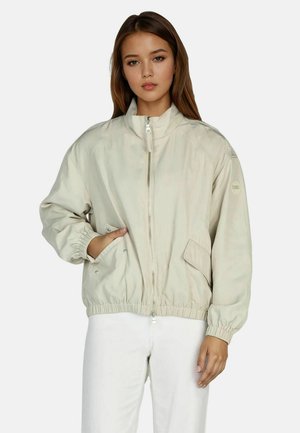 Young woman with light brown hair wearing a beige zip-up jacket with elastic cuffs and white pants against a plain background.