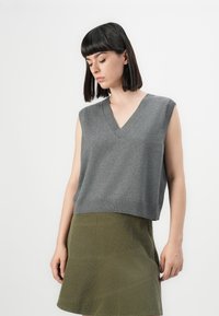Woman with straight black hair wears sleeveless gray V-neck sweater and olive green skirt, standing against plain white background.