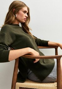 Maglione a maglia verde oliva con polsini a costine e collo alto, che cade su pantaloni corti grigi a fantasia. Modella seduta su una sedia di legno.
