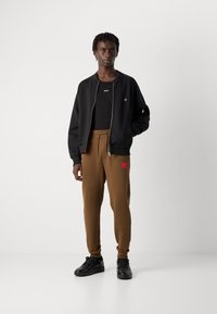 Young man standing wearing black zip-up jacket, black shirt, brown drawstring pants with red patch, and black sneakers against plain white background.