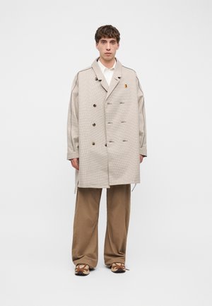 Giovane uomo che indossa un cappotto beige doppiopetto oversize, camicia bianca, pantaloni marroni larghi e sneakers marroni con motivi, in piedi su sfondo bianco.