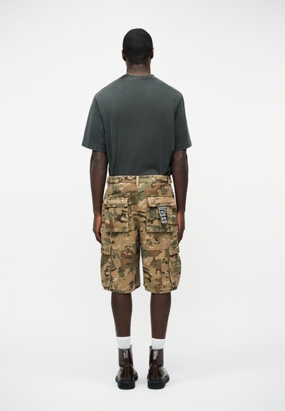 Άνδρας με σκουρόγρινη μπλούζα, καμουφλαρισμένες cargo βερμούδες με πολλές τσέπες και μαύρες μπότες, στέκεται με την πλάτη προς τα εμπρός σε απλό λευκό φόντο.