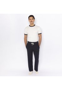 White T-shirt with black trim and matching navy sweatpants with drawstring. Model stands casually with hands in pockets, wearing light-colored shoes.