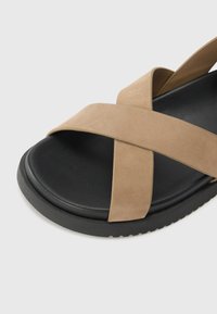 Sandalia beige con tiras cruzadas, de textura suave y suela negra. Cuenta con una plantilla cómoda y forma redondeada. Sin herrajes visibles.