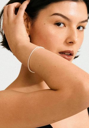 Silver bracelet with small clear stones, worn on a light brown arm. Smooth skin texture, short dark hair framing the face.