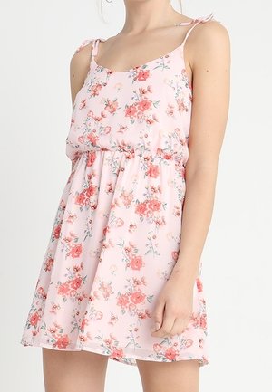 Femme portant une robe rose sans manches avec des motifs floraux rouges et blancs, des bretelles nouées et une taille froncée.