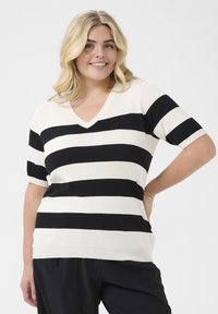 Pulover cu mânecă scurtă și decolteu în V, de culoare crem cu dungi negre îndrăznețe. Textură tricotată cu o croială relaxată. Accesoriu de bijuterie vizibil pe încheietura mâinii.