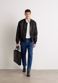 Chaqueta bomber negra sobre una camiseta blanca, combinada con jeans azules y botas negras hasta el tobillo. Lleva un bolso tote negro. Tejido suave y corte casual.