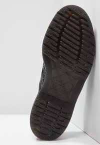 Semelle d'une botte noire montrant des motifs de sculpture profonds, des zones de grip texturées et un logo rectangulaire avec du texte et des symboles au centre.