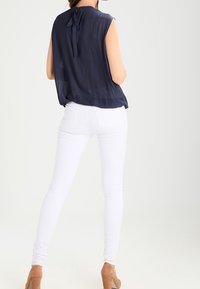 Kvinna iklädd ärmlös marinblå blus med knytbak, vita figurnära byxor och tanfärgade högklackade skor, stående med ryggen mot kameran.