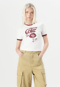 White short-sleeve t-shirt featuring a red "cherry Coke" graphic print and maroon ribbed neckline. Paired with beige cargo pants.