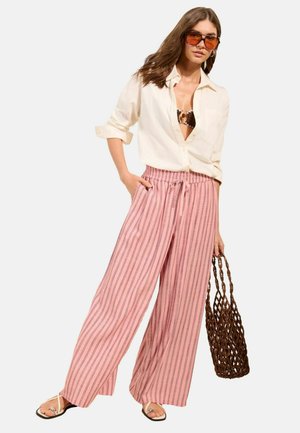 Kvinde iført oversized cremefarvet skjorte over mønstret bikini-top, lyserøde stribede vidde bukser, sandaler, brun net-taske og orange solbriller.